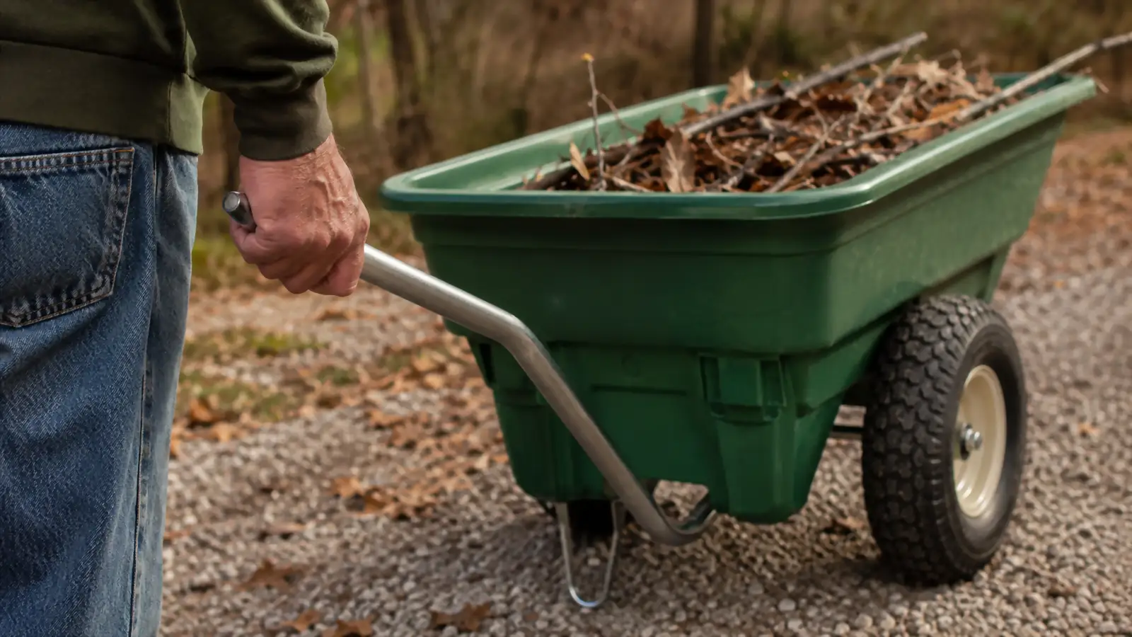 Close-up of a hand gripping the pull handle of a green poly dump cart, the bed loaded with dead leaves and small branches.